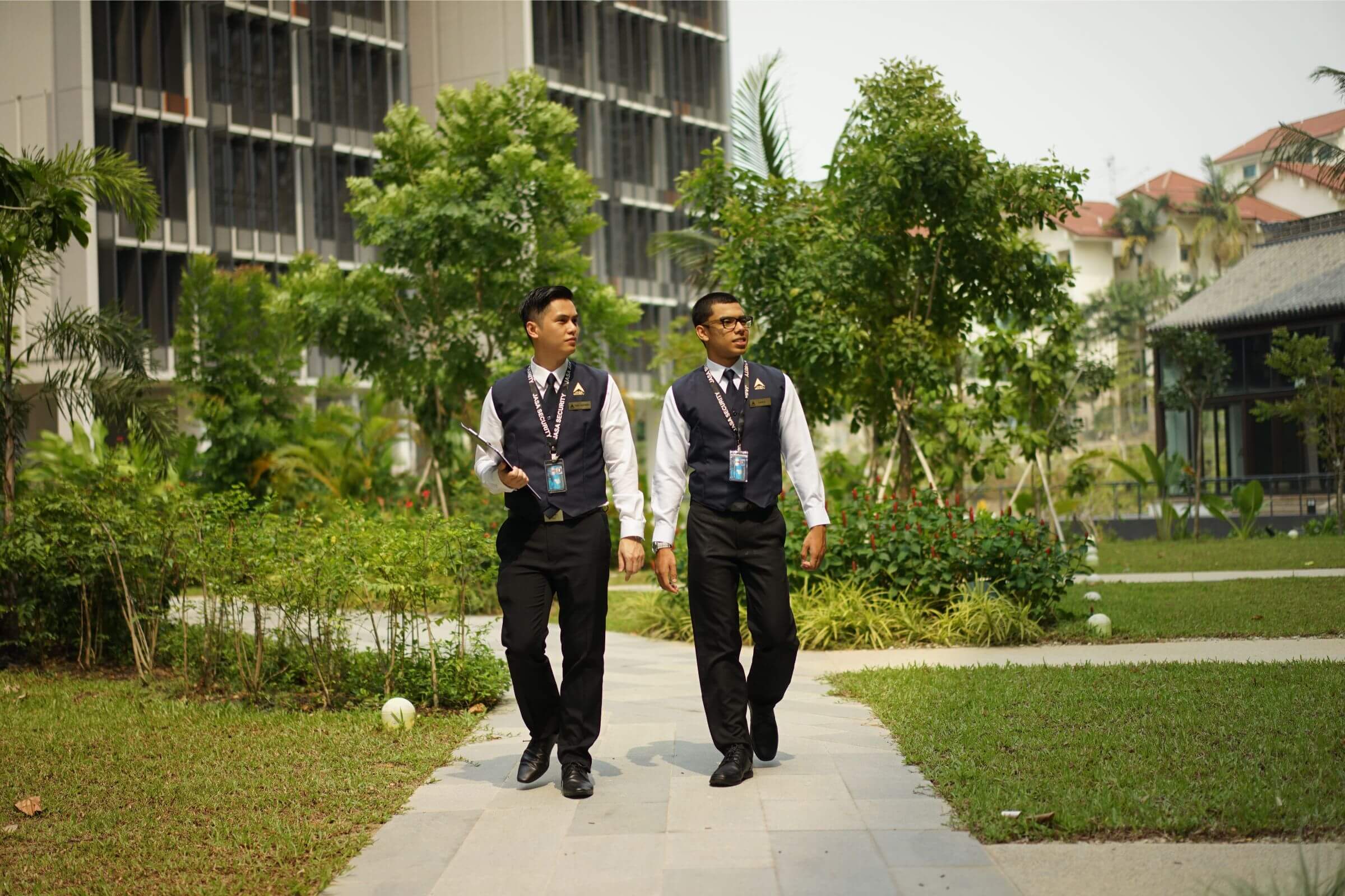 Two uniformed Jasa Security officers conducting outdoor patrol in a landscaped residential or commercial complex, demonstrating professionalism and vigilance